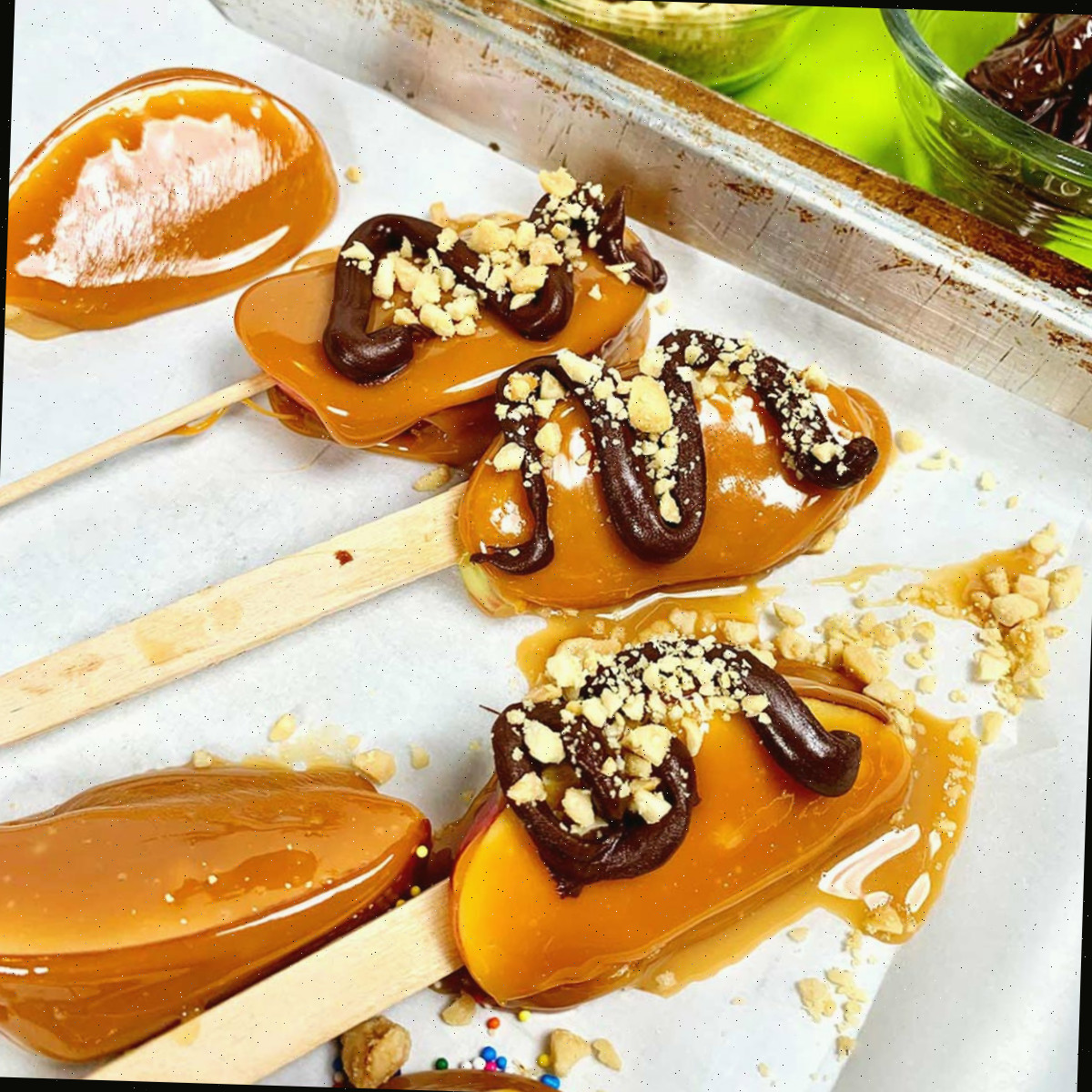 Fatias de maçã caramelizada completas em palitos descansando em papel manteiga. Algumas têm espirais de chocolate meio amargo e nozes polvilhadas.