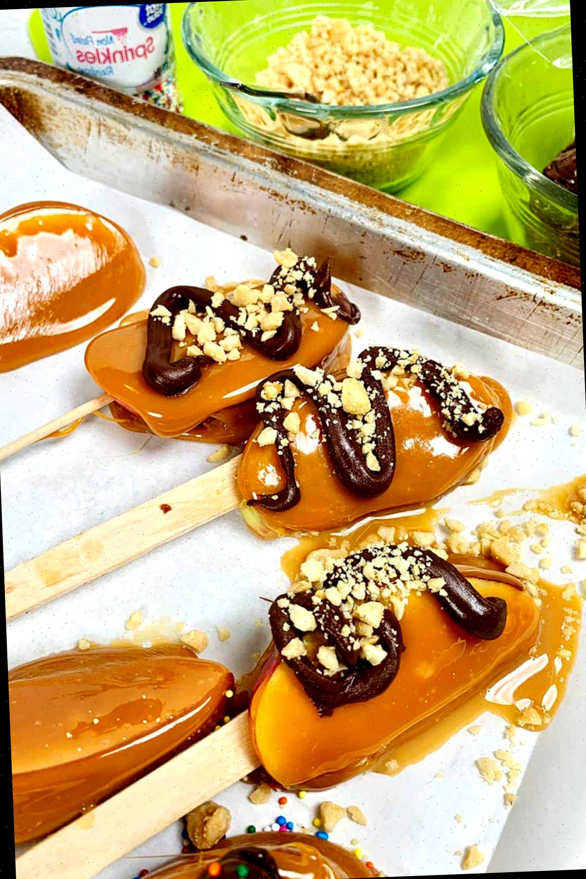 Fatias de maçã caramelada concluídas no palito repousando sobre papel manteiga. Algumas têm espirais de chocolate amargo e nozes polvilhadas.