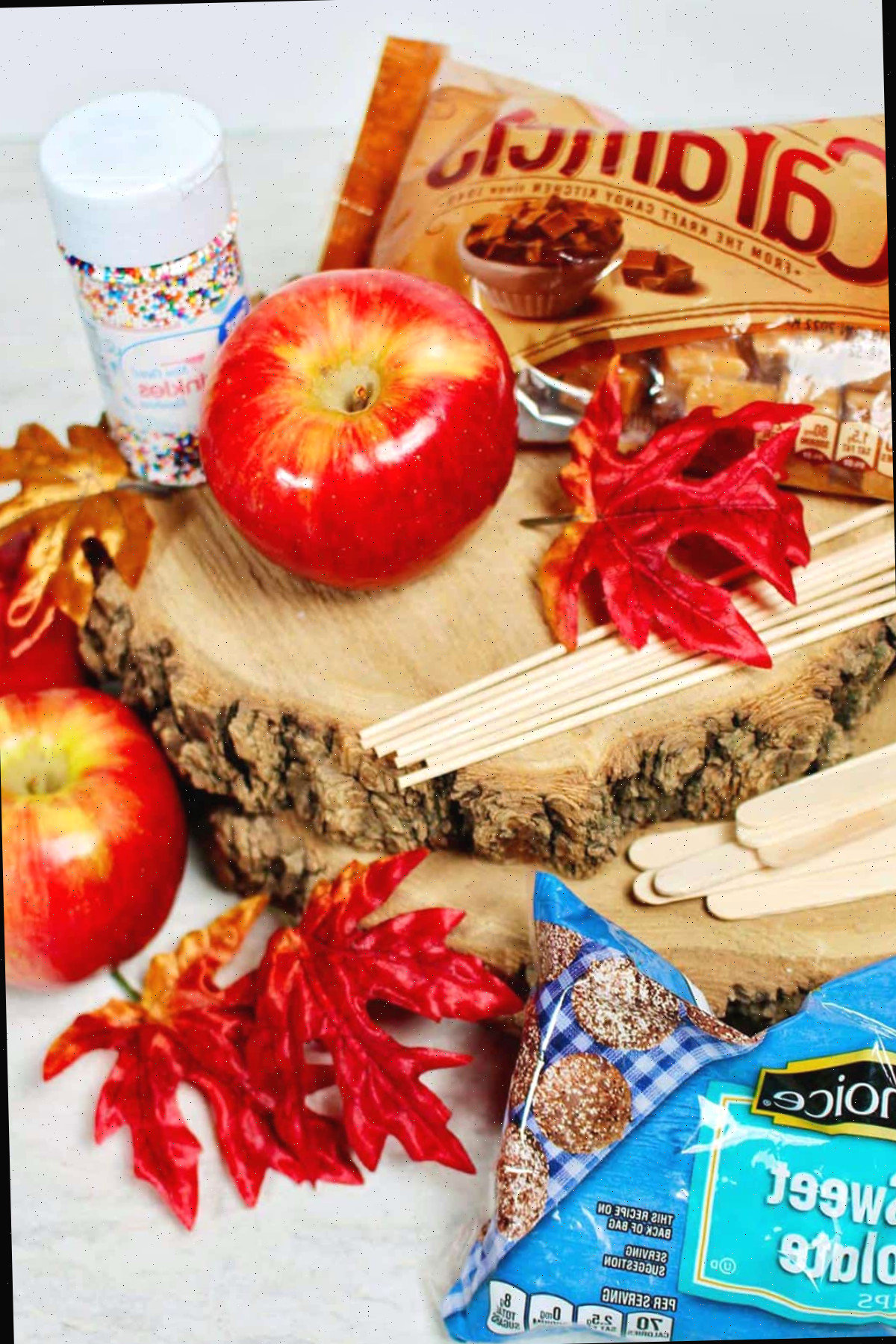 Exibição da temporada de outono com maçãs vermelhas, palitos, gotas de chocolate meio amargo, caramelos, granulados e folhas sobre fatias de madeira.