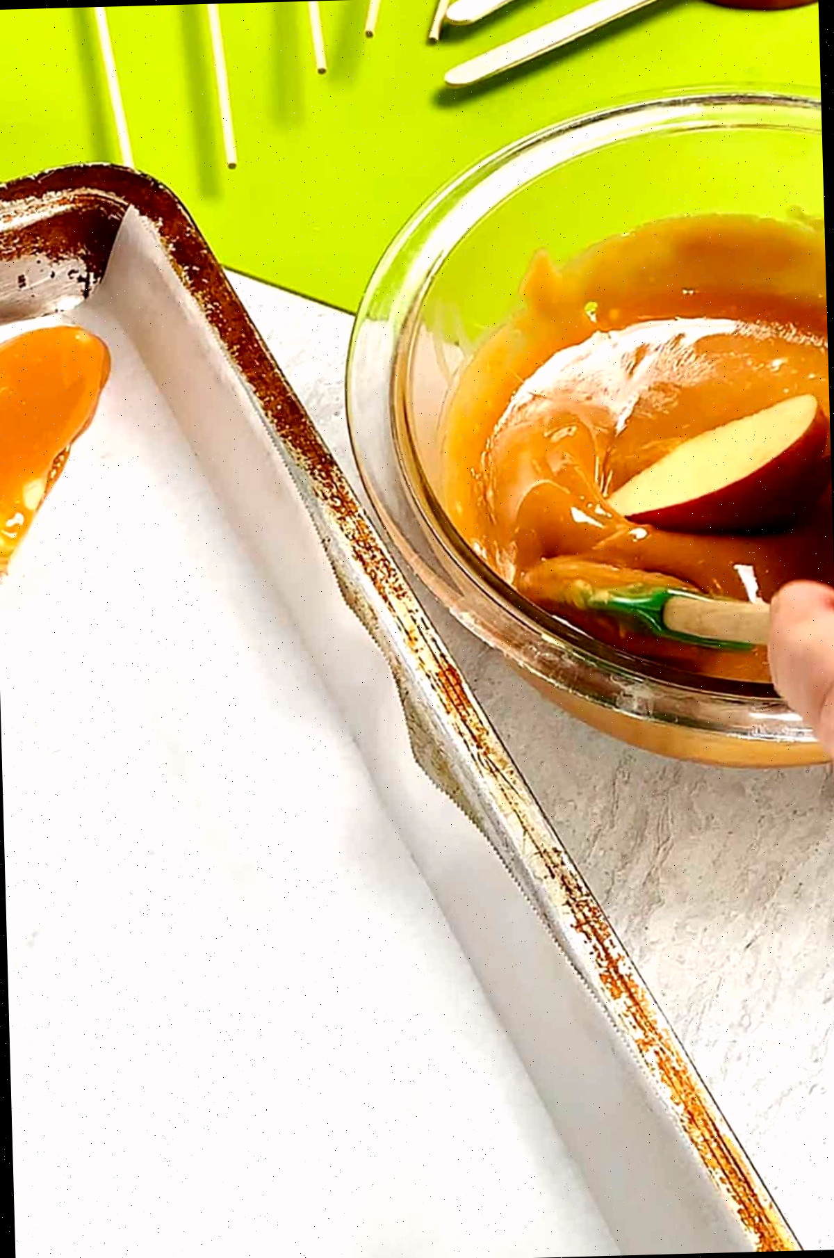 Pessoa mergulhando fatias de maçã no palito em caramelo aquecido em uma tigela ao lado de uma tábua de cortar verde brilhante e uma assadeira com papel manteiga.