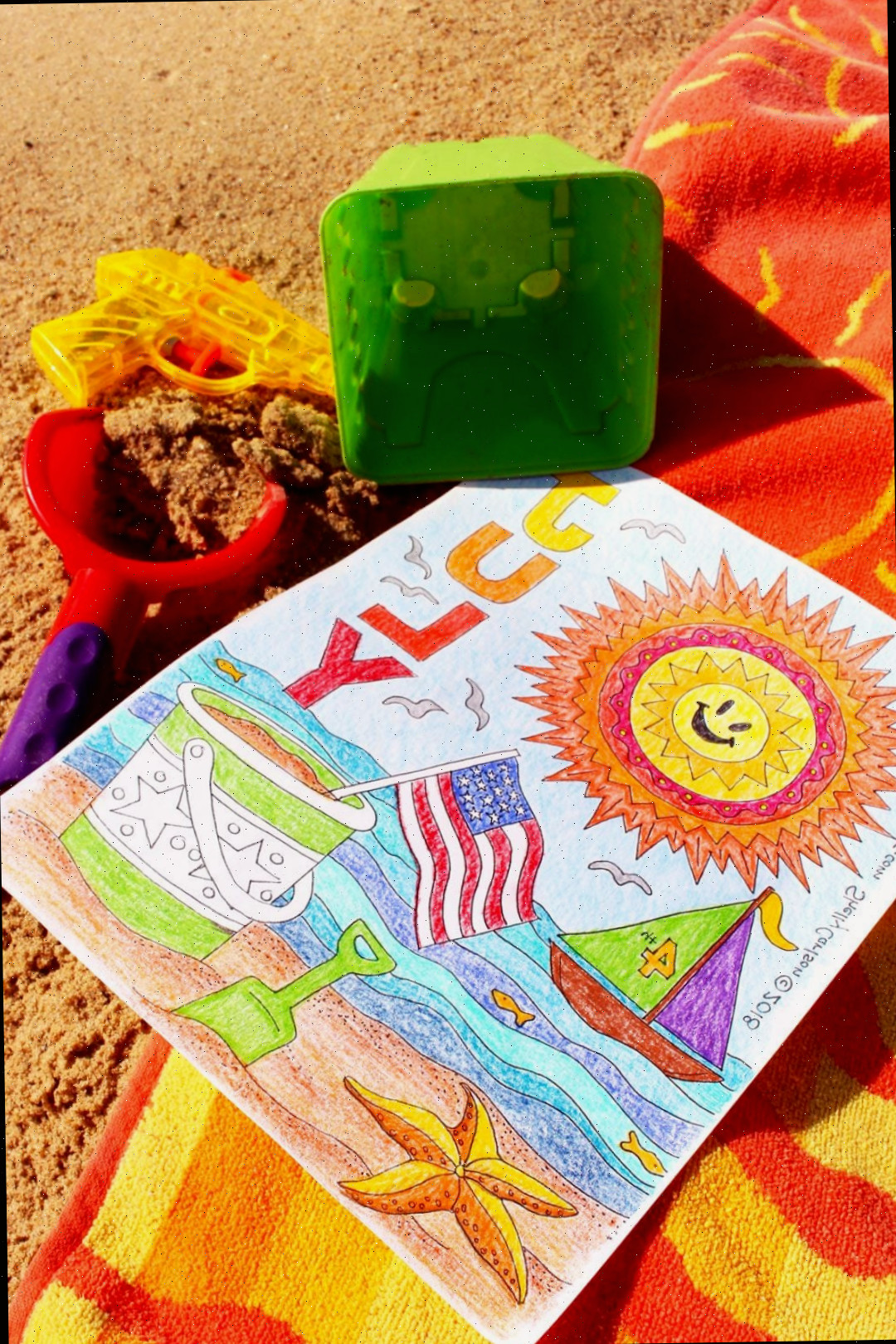Página de colorir de julho completada em toalha de praia na areia com lápis de cor ao redor.