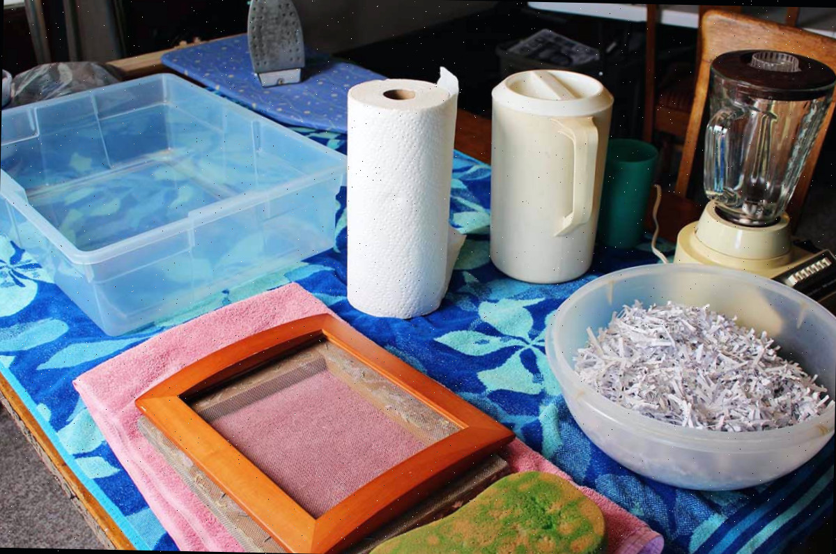 Uma tigela de aparas de papel, uma moldura de quadro com molde e deckle, toalhas de papel, uma toalha de praia, um recipiente com água, uma imagem e um ferro.