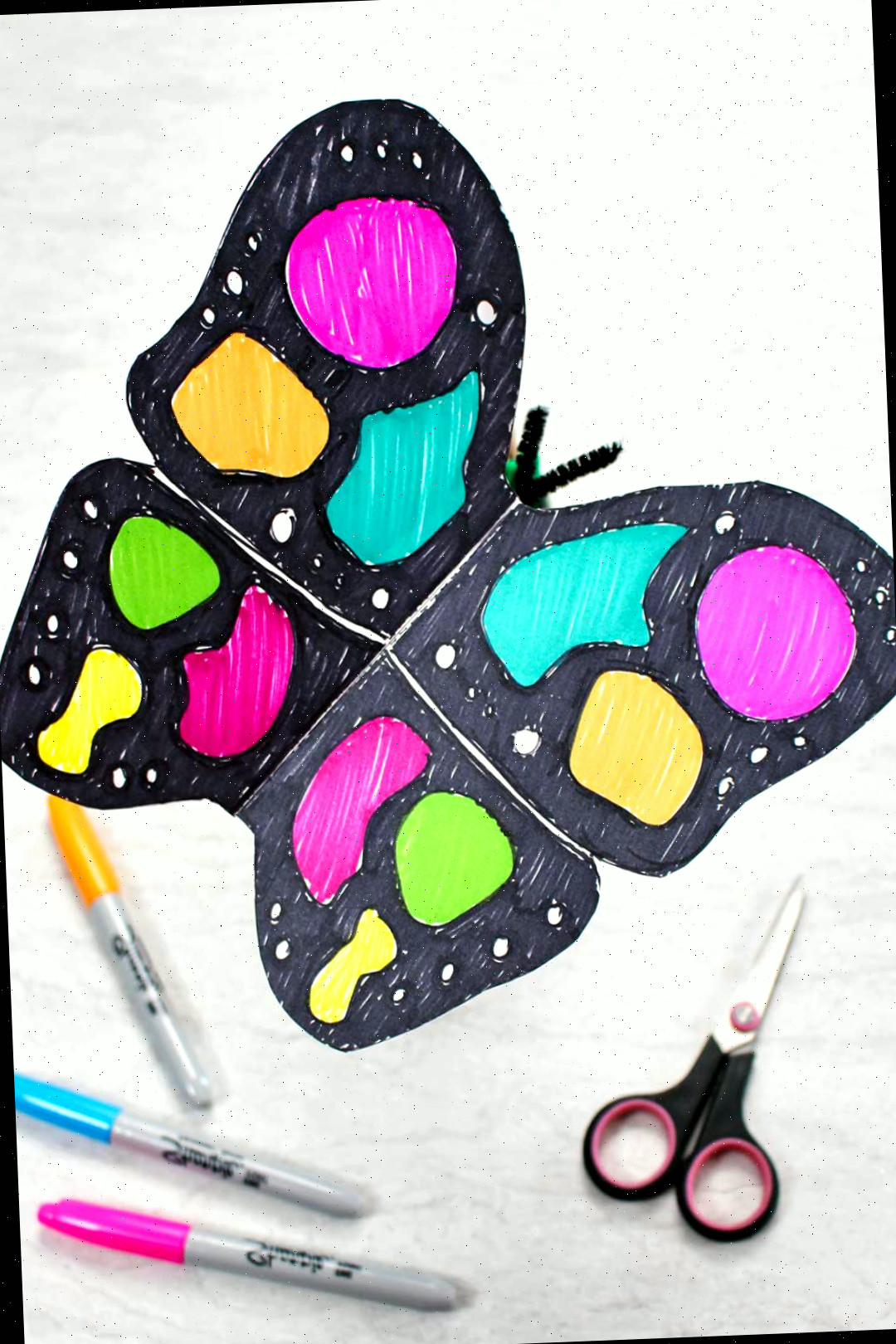 Artesanato de borboleta de papel vibrante com marcadores pretos e neon repousando próximo a uma tesoura preta e marcadores neon.