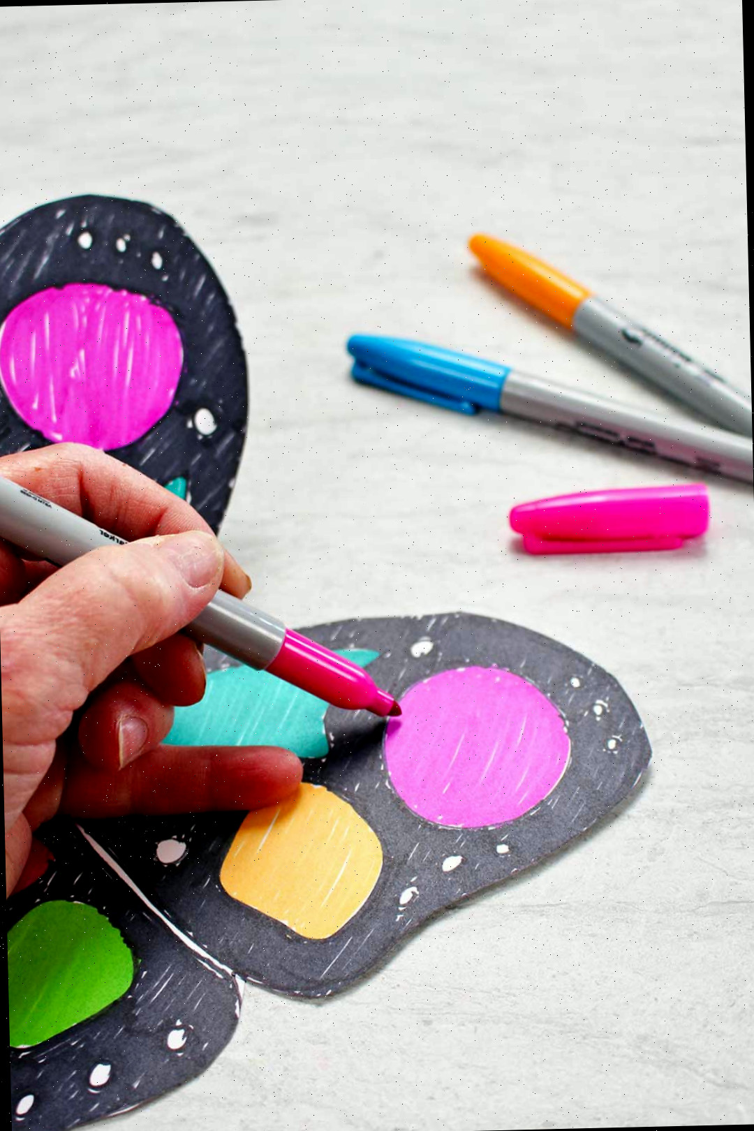 A mão de uma pessoa colorindo uma parte da asa de papel da borboleta com um marcador neon rosa com outros marcadores descansando próximos.