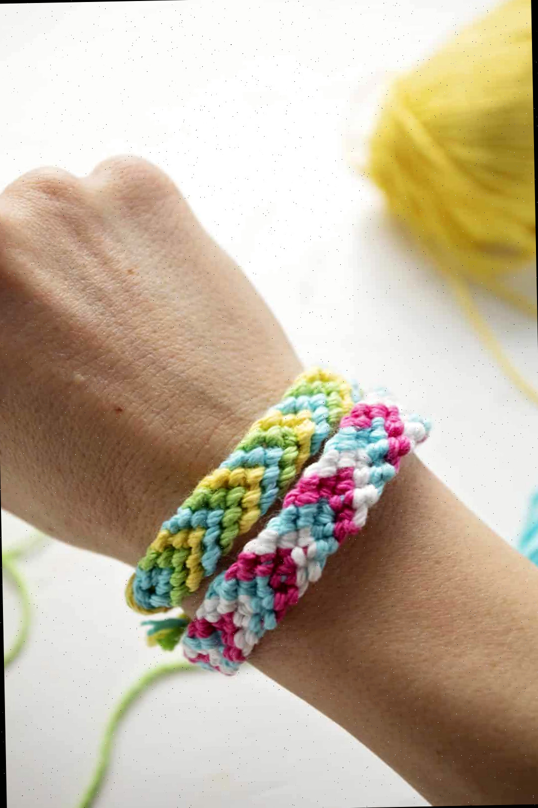Duas pulseiras de padrão chevron em fio de bordado verde, azul, amarelo, rosa e branco, amarradas no pulso de uma pessoa.