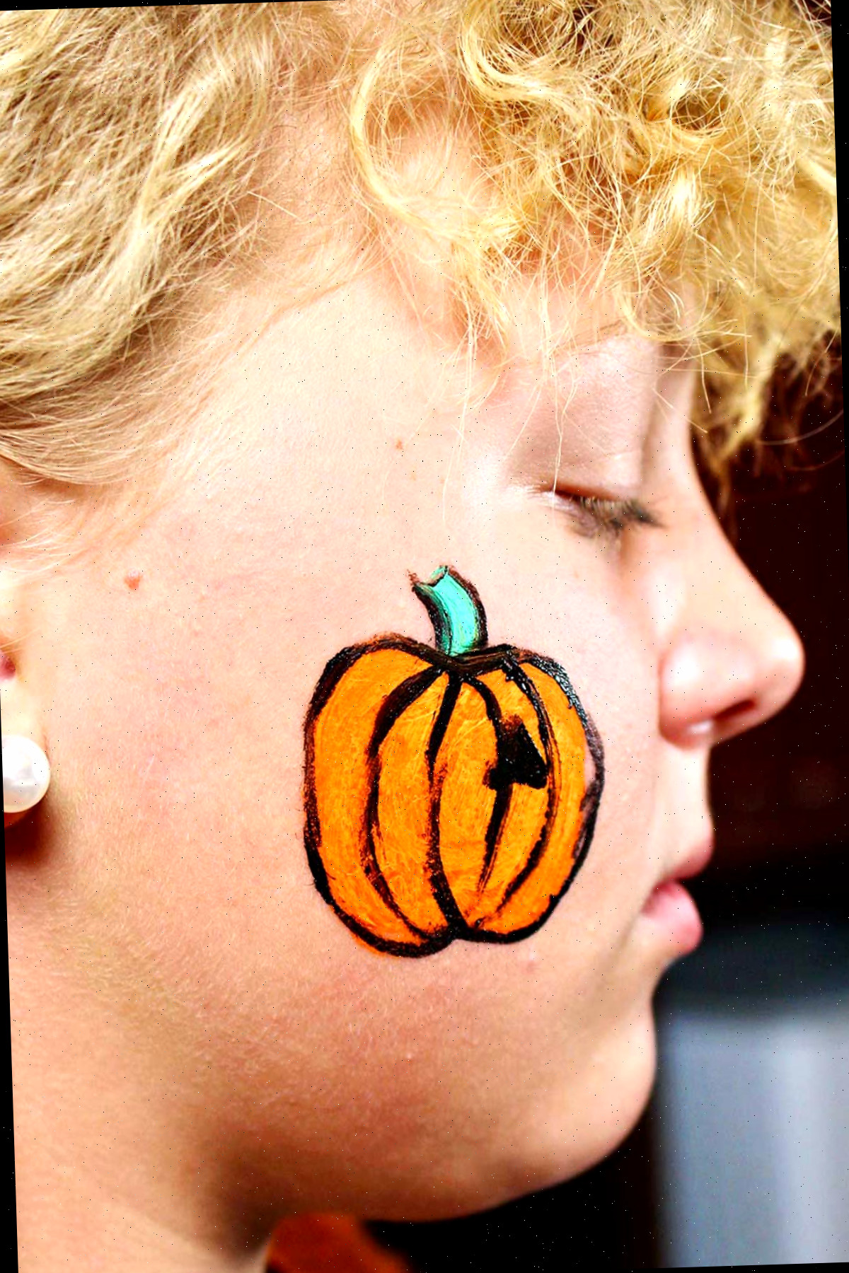 Perfil do rosto de uma menina com pintura mais detalhada de uma abóbora laranja com talo verde e contorno preto.