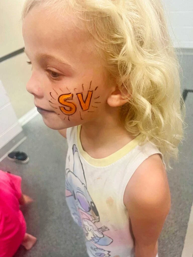 Menina jovem com cabelo loiro e pintura facial laranja "SV".