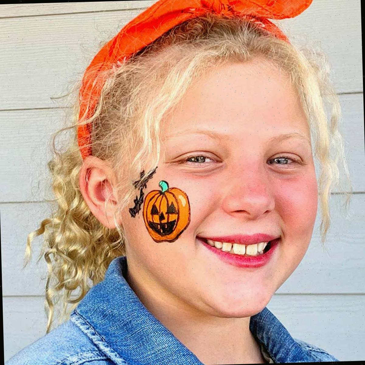 Menina com cabelo loiro encaracolado sorrindo para a câmera com pintura de rosto de abóbora de Halloween na bochecha.