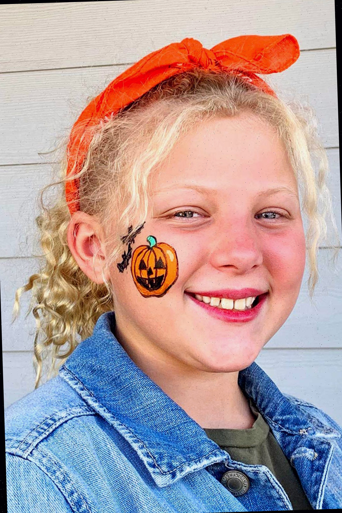 Menina com cabelo encaracolado loiro sorrindo para a câmera com pintura de rosto de abóbora de Halloween na bochecha.