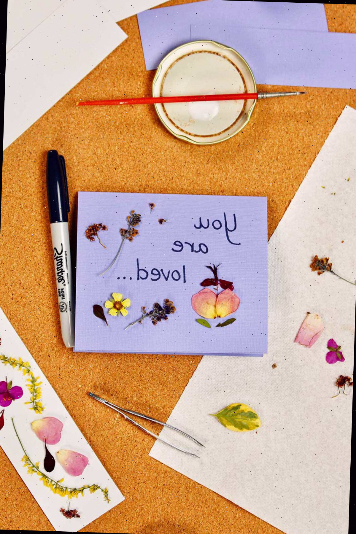 Uma carta roxa dizendo 'Você é querido...' com flores prensadas sobre ela, além de um marcador também com flores prensadas.