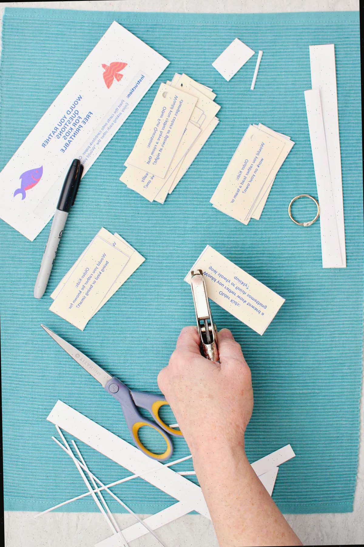 Alguém furando um buraco em uma pergunta de Would You Rather em um pequeno cartão, com pedaços de papel cortados, uma tesoura, um furador de papel e uma caneta próximos.