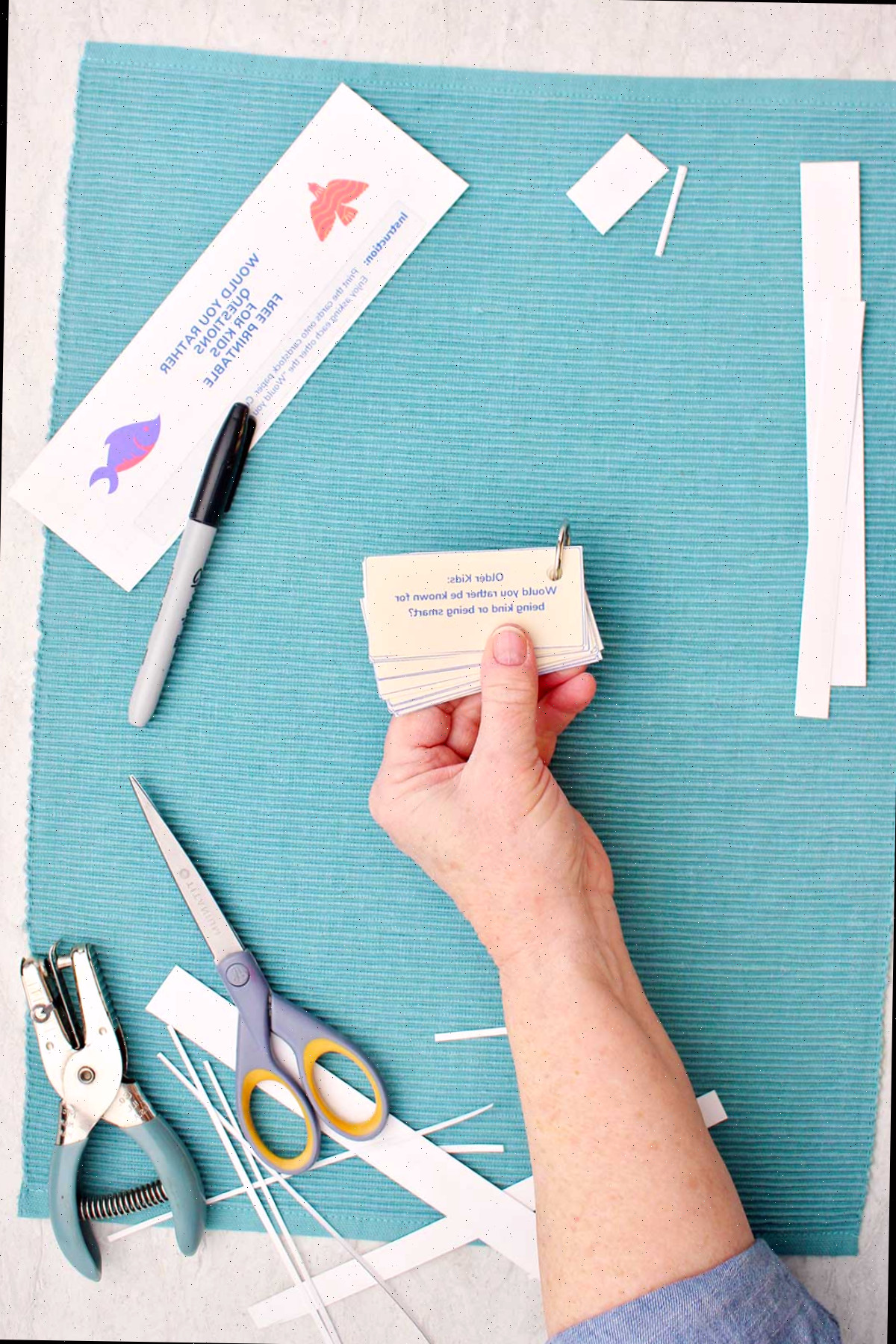 Alguém segurando uma pilha de perguntas Você Preferiria com pedaços de papel cortados, uma tesoura, um furador e um marcador ao lado.