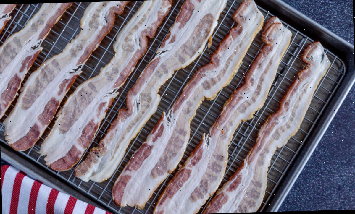 Fatias de bacon em uma grade de arame sobre uma assadeira ao lado de uma toalha vermelha e branca.
