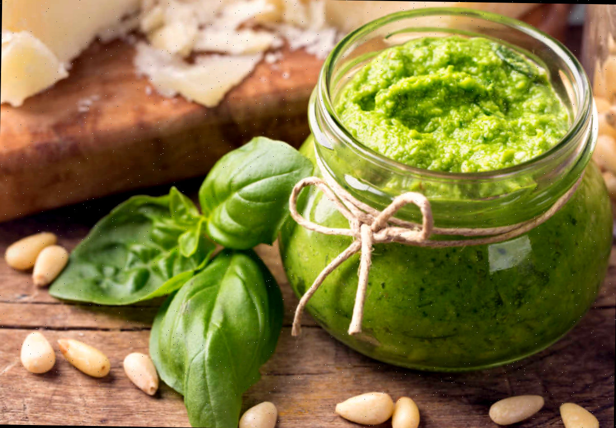 Pesto de manjericão em um recipiente de vidro sobre uma tábua de madeira, ao lado de manjericão fresco, pinhões e queijo parmesão.