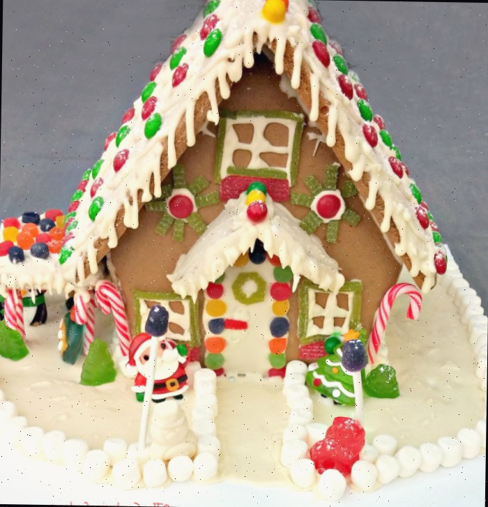 Residência de gengibre com telhado coberto de glacê e enfeites de guloseimas, cercada por um jardim de doces contornado com marshmallows.