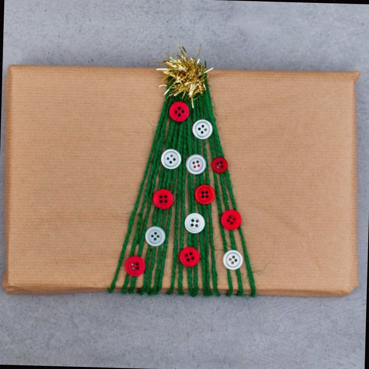 Uma ideia criativa de embalagem de presente de férias mostrando um pacote retangular embrulhado em papel marrom, com fio verde envolvendo-o para se parecer com uma árvore, botões colados para representar enfeites e um pequeno pedaço de guirlanda no topo para se parecer com uma estrela de topo de árvore.