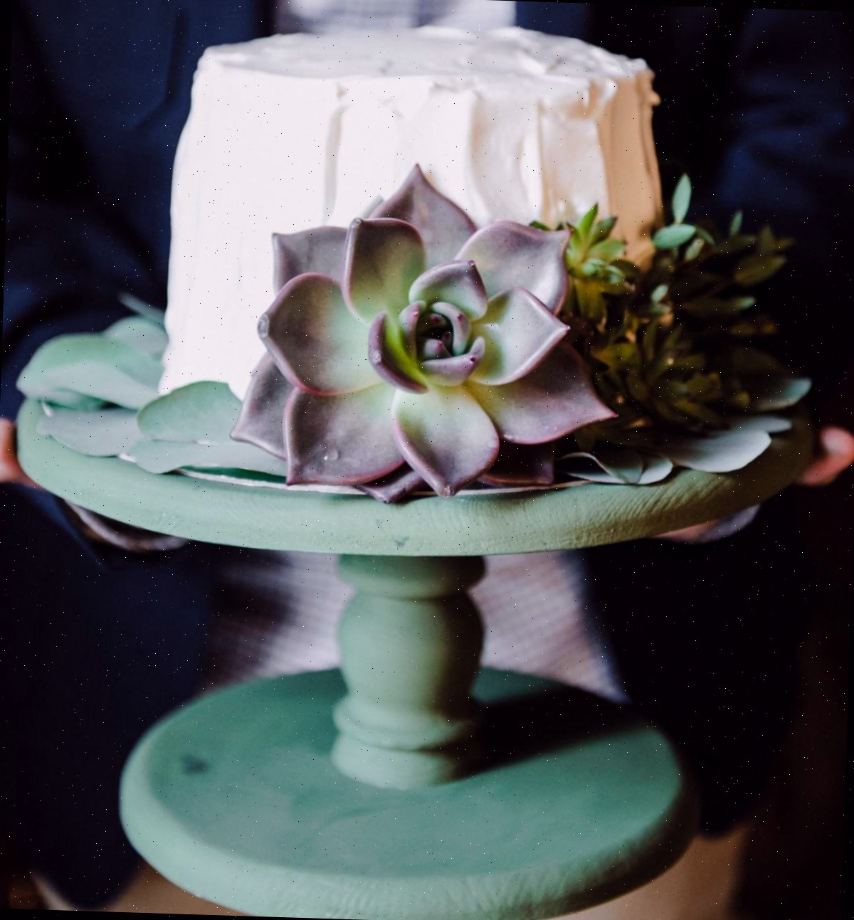 Bolo com suculentas em suporte de bolo verde Um bolo com suculentas coberto com buttercream branca em um pedestal de madeira verde, com suculentas de pasta de goma ao redor do bolo.