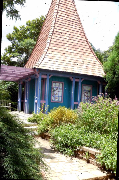 Pequena casa azul com telhado curvo, cercada por bordas exuberantes e um caminho.