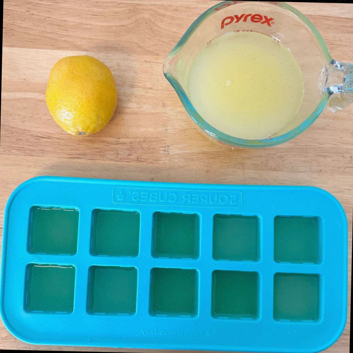 Congelando suco de limão Uma imagem do processo de congelar suco de limão em bandejas de gelo de silicone, que mostra um recipiente Pyrex de uma xícara cheio de suco de limão ao lado de um limão, acima de uma bandeja de gelo azul reutilizável cheia de suco de limão.