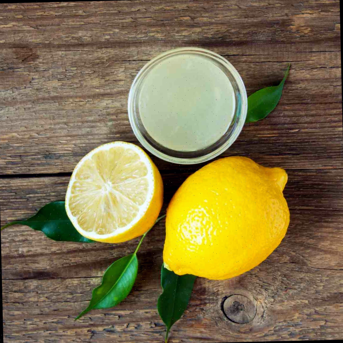 Um ramequim de vidro com suco de limão recém espremido acima de um limão inteiro e um limão cortado ao meio com várias folhas de limão ao redor.