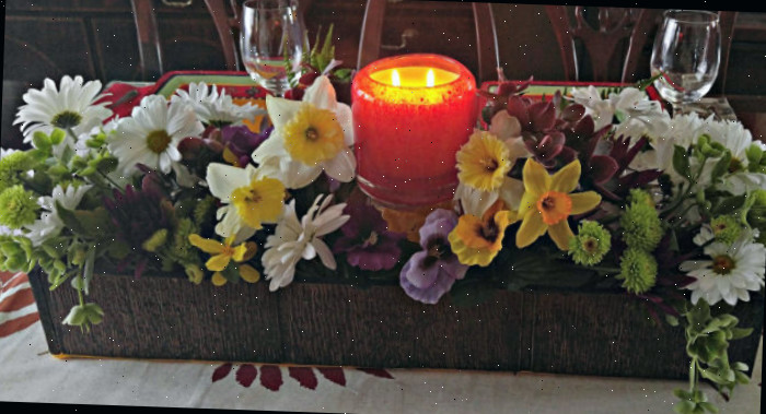Uma caixa de madeira marrom repleta de flores artificiais e uma vela acesa em um recipiente de vidro cria um lindo arranjo central.