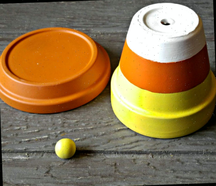 Vaso de barro pintado de laranja, amarelo e branco, saucer de barro pintado de laranja e uma pequena bola de madeira pintada de amarelo em cima de uma mesa de madeira.