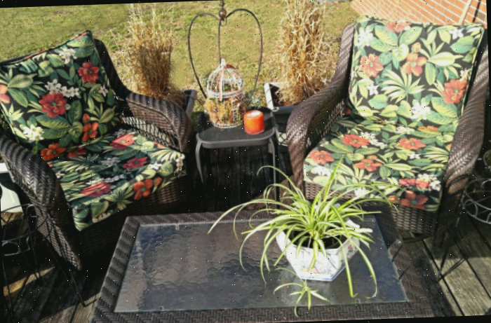 Um espaço de pátio com duas cadeiras e almofadas, uma planta e uma mesa pequena decorada com uma vela de vidro.