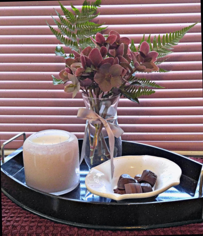 Heléboros e samambaias em um vaso de vidro, ao lado de uma tigela de chocolate e uma vela de vidro. Uma cortina rosa fechada e um corredor de mesa adicionam calor à cena.