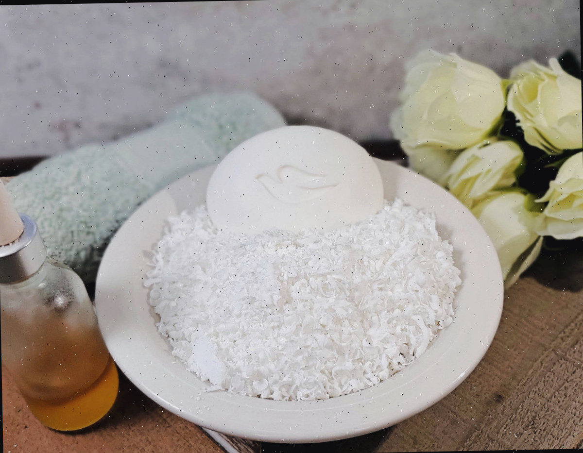 Rale as lascas de sabão Dove Uma tigela branca com lascas de sabão Dove e uma barra de sabão ao lado de rosas brancas, uma toalha verde enrolada e um frasco de óleo essencial.