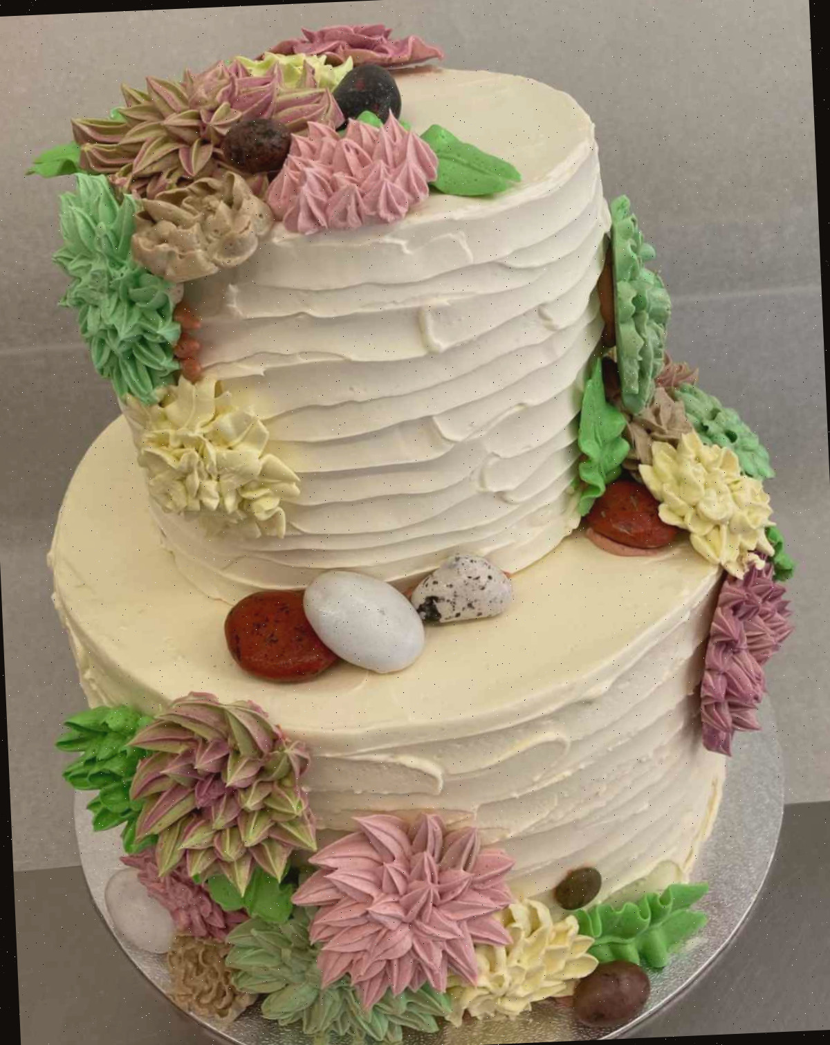 Um design de bolo com suculentas de buttercream Um bolo com suculentas de buttercream com duas camadas, coberto com cobertura branca, apresentando suculentas de buttercream (modeladas após echeveria) em rosa, amarelo, verde e marrom descendo pelas camadas do bolo.