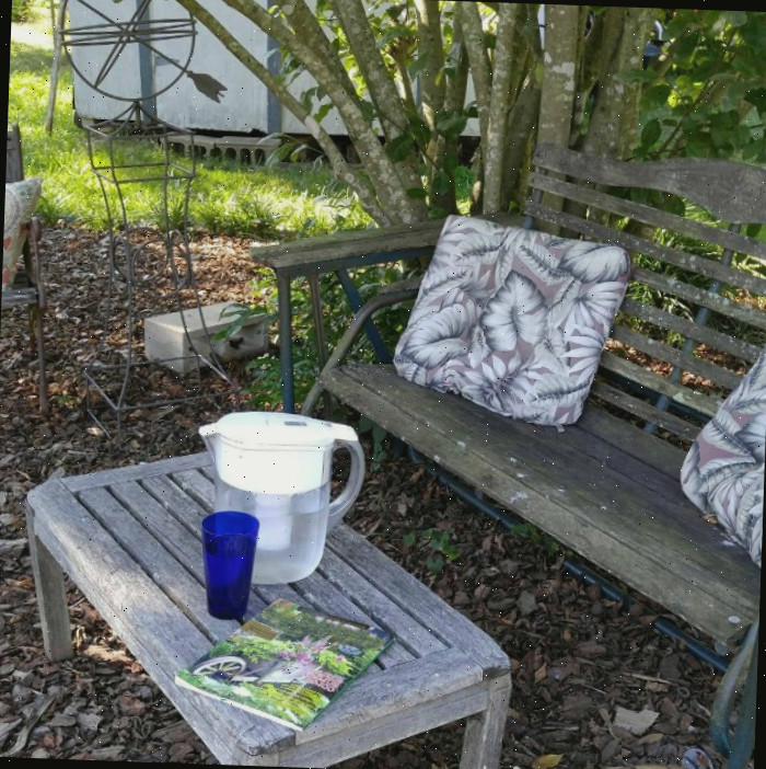 Ambiente de jardim com almofadas, jarra de água, copo e revista em uma mesa de madeira.